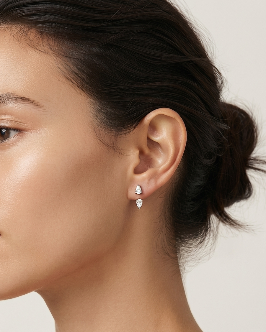 A close-up side profile of a woman wearing a pair of small PEAR DIAMOND earrings with clear stones, set against a clean, light beige background.