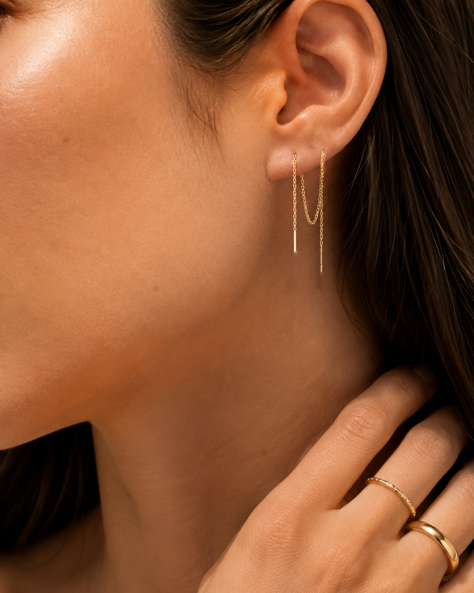 A close-up of a model's ear wearing a delicate gold double-drop chain earring, with her hand resting near her neck and two gold rings on her fingers.