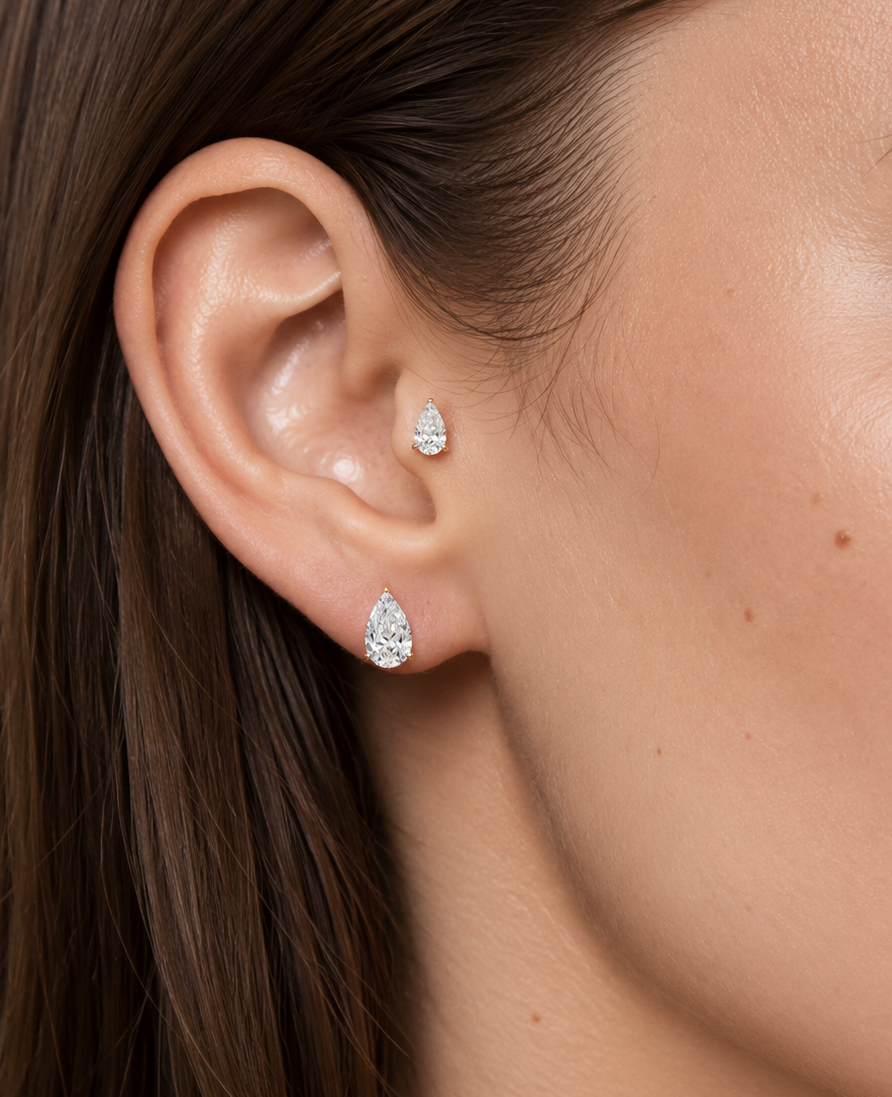 Close-up of a woman's ear wearing two small pear-shaped stud earrings with clear sparkling stones.