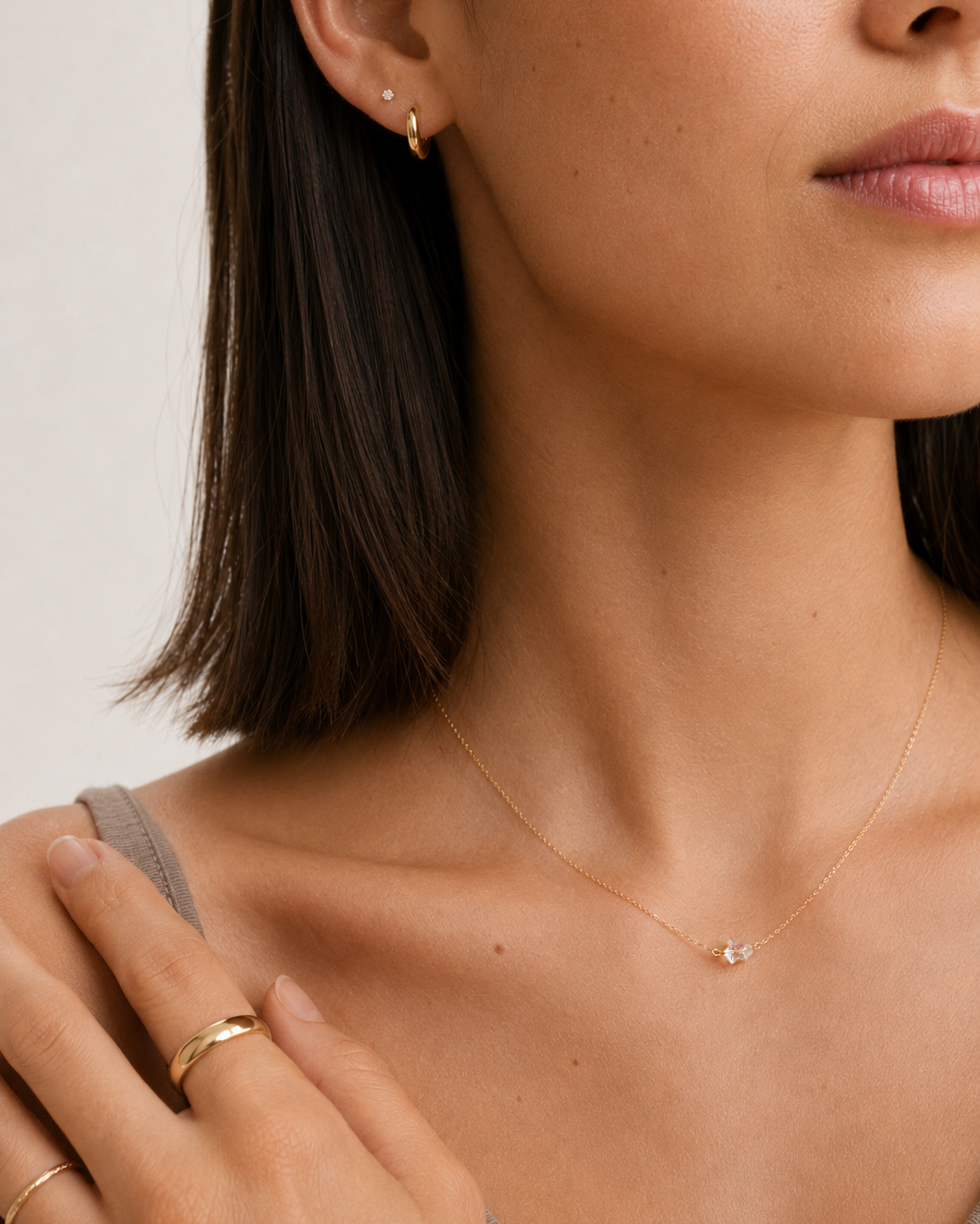 Close-up of a woman wearing a delicate gold chain necklace with a small clear stone pendant, along with matching gold rings and a small gold hoop earring.