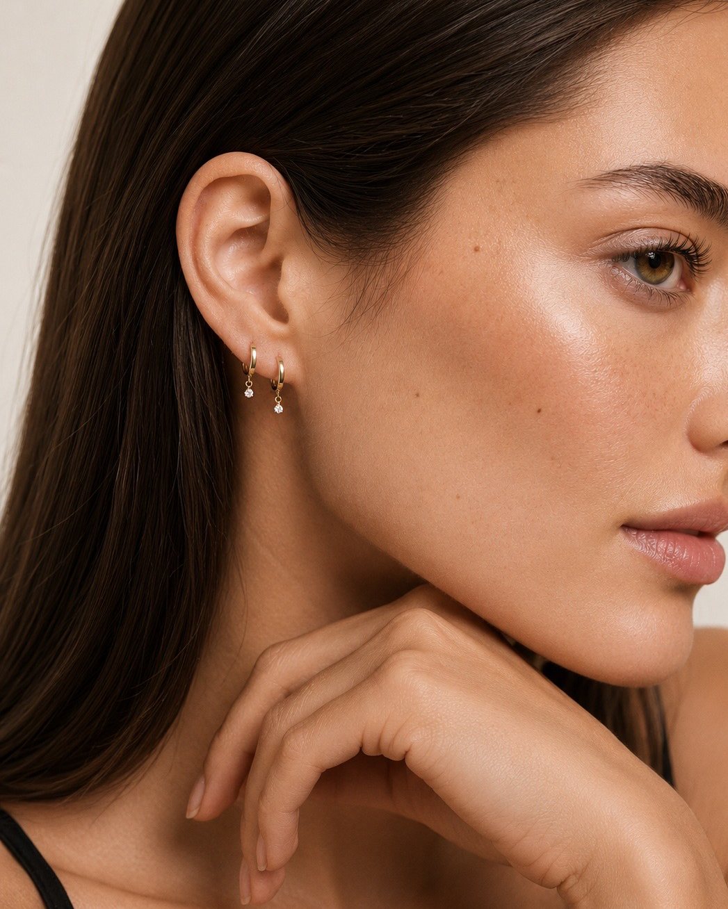 A close-up of a woman wearing a delicate gold huggie hoop earring with small dangling teardrop crystals, shown against a soft beige background.