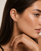 A close-up of a woman wearing a delicate gold huggie hoop earring with small dangling teardrop crystals, shown against a soft beige background.