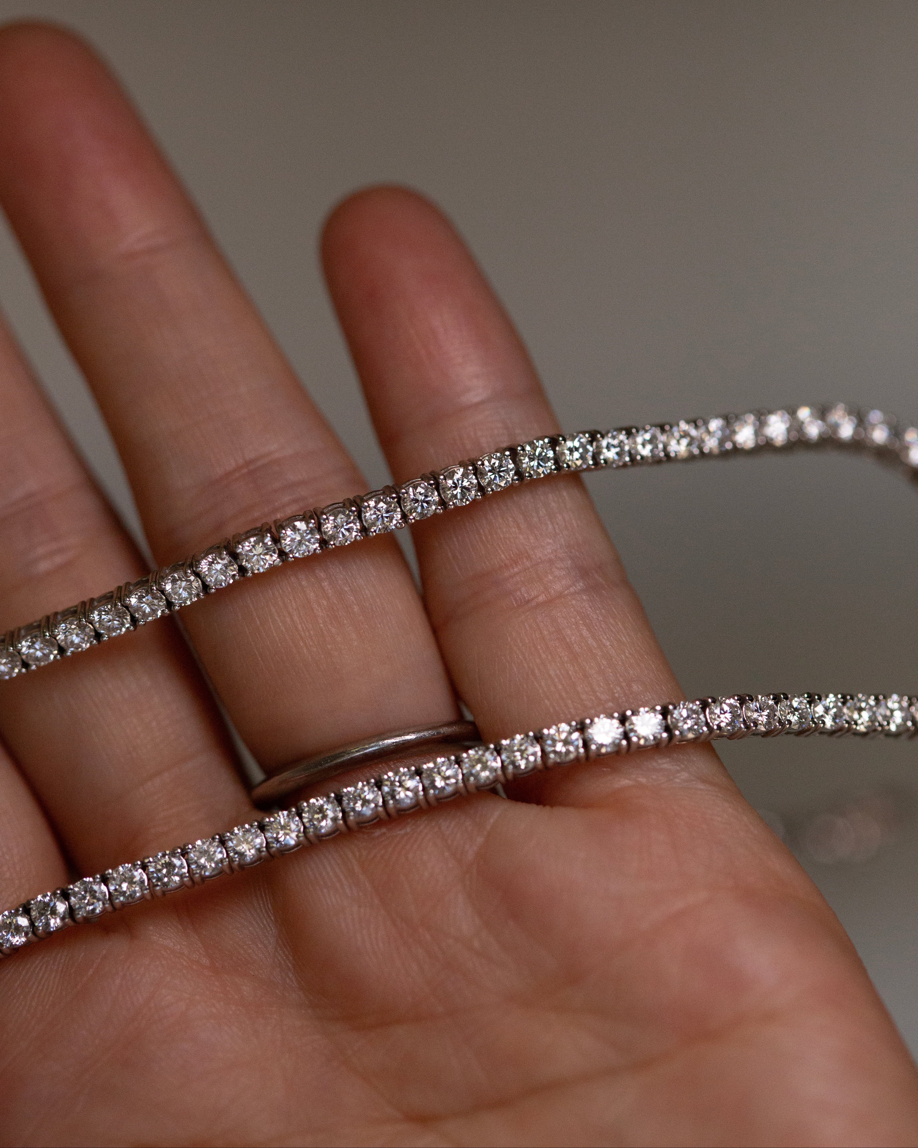 A close-up of a hand holding a delicate white gold tennis bracelet with brilliant diamonds, with a matching silver band visible on the ring finger.