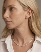 A close-up side profile of a blonde woman wearing a delicate silver tennis necklace and a white collared shirt against a soft beige background.