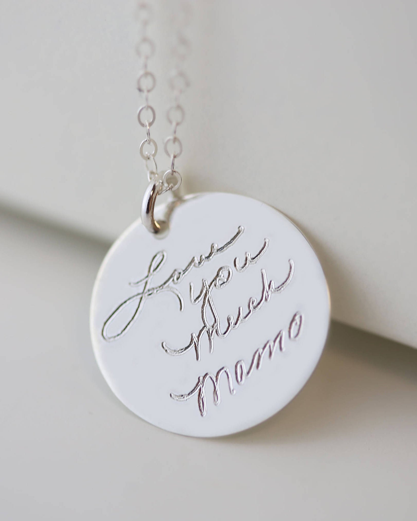 A silver necklace with a round pendant that says 'Love you much Mama' in cursive handwriting.